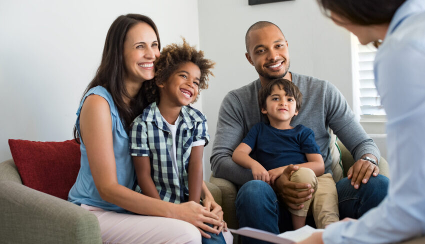 Happy young family sitting on couch and talking with family counselor. Smiling parents with adopted children discussing with counselor. Multiethnic family meeting a financial agent.