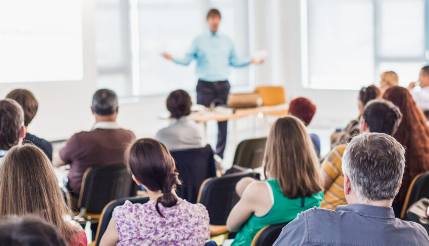 Business and entrepreneurship symposium. Speaker giving a talk at business meeting. Audience in conference hall. Rear view of unrecognized participant in audience.